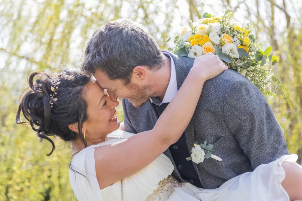 Jeune couple de mariés s'embrassant tendrement sous une pergola ornée de roses, dans un cadre bucolique.