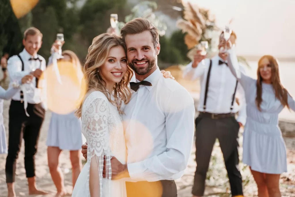 Jeune couple se tenant par la main, échangeant un regard tendre lors d'une réception de mariage, avec des invités et des décorations élégantes en arrière-plan